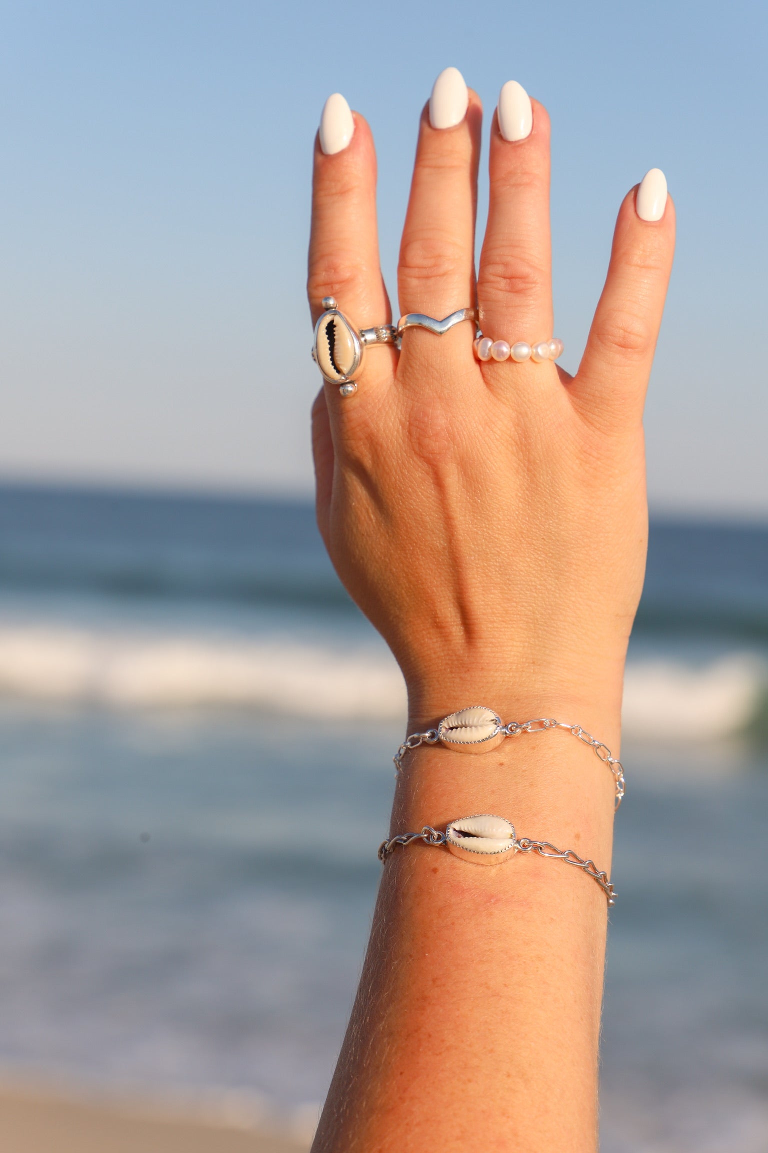 A close-up of a person's hand with white manicured nails, wearing silver shell and pearl jewelry, including two silver bracelets and three rings, set against a blurred beach background.