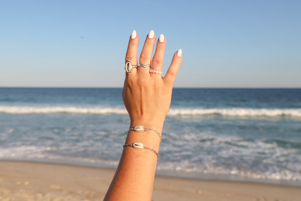 A hand with white manicured nails and silver rings and bracelets, raised against a blurred beach and ocean background.